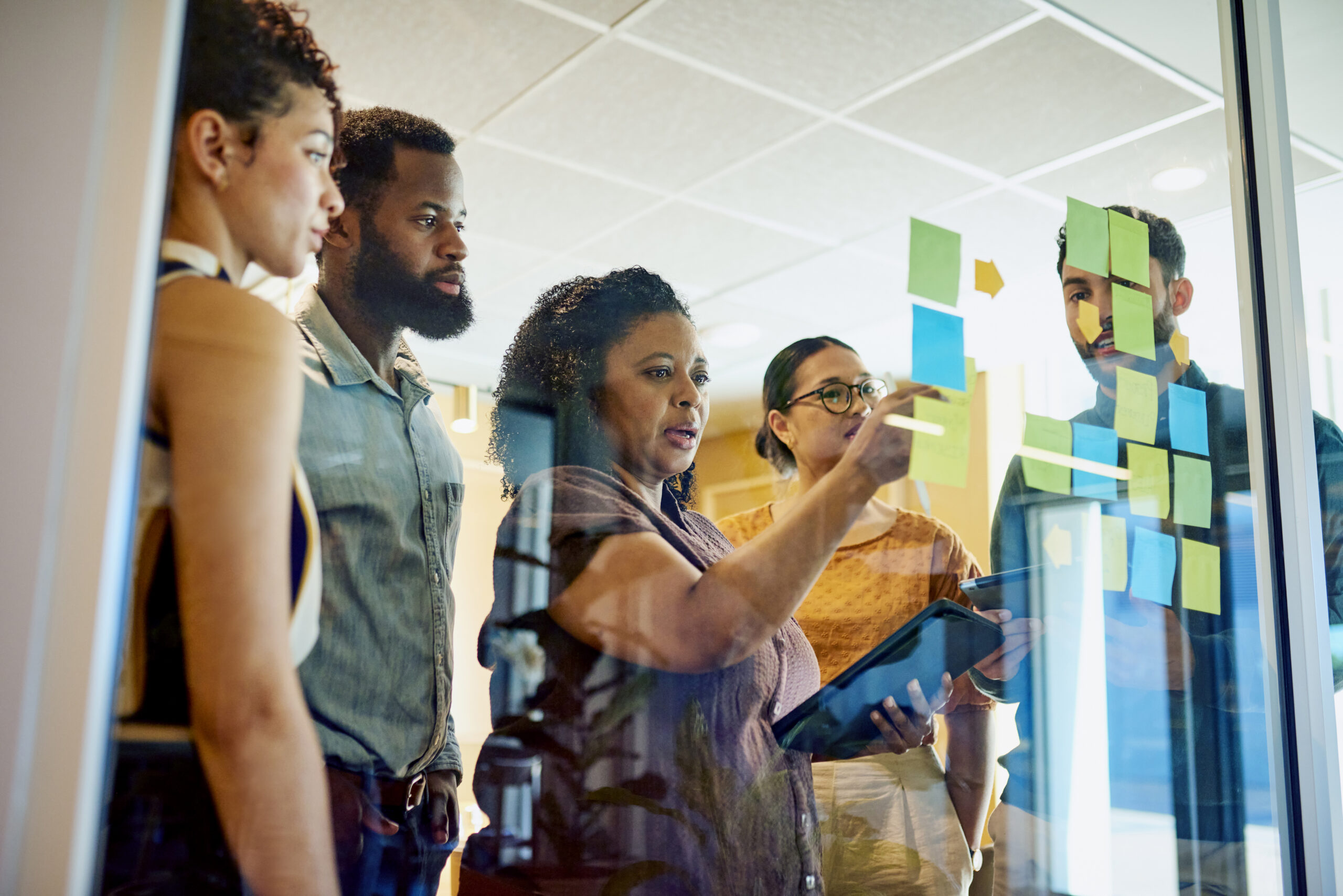 A group of diverse professionals are brainstorming ideas while standing around a glass wall with sticky notes.